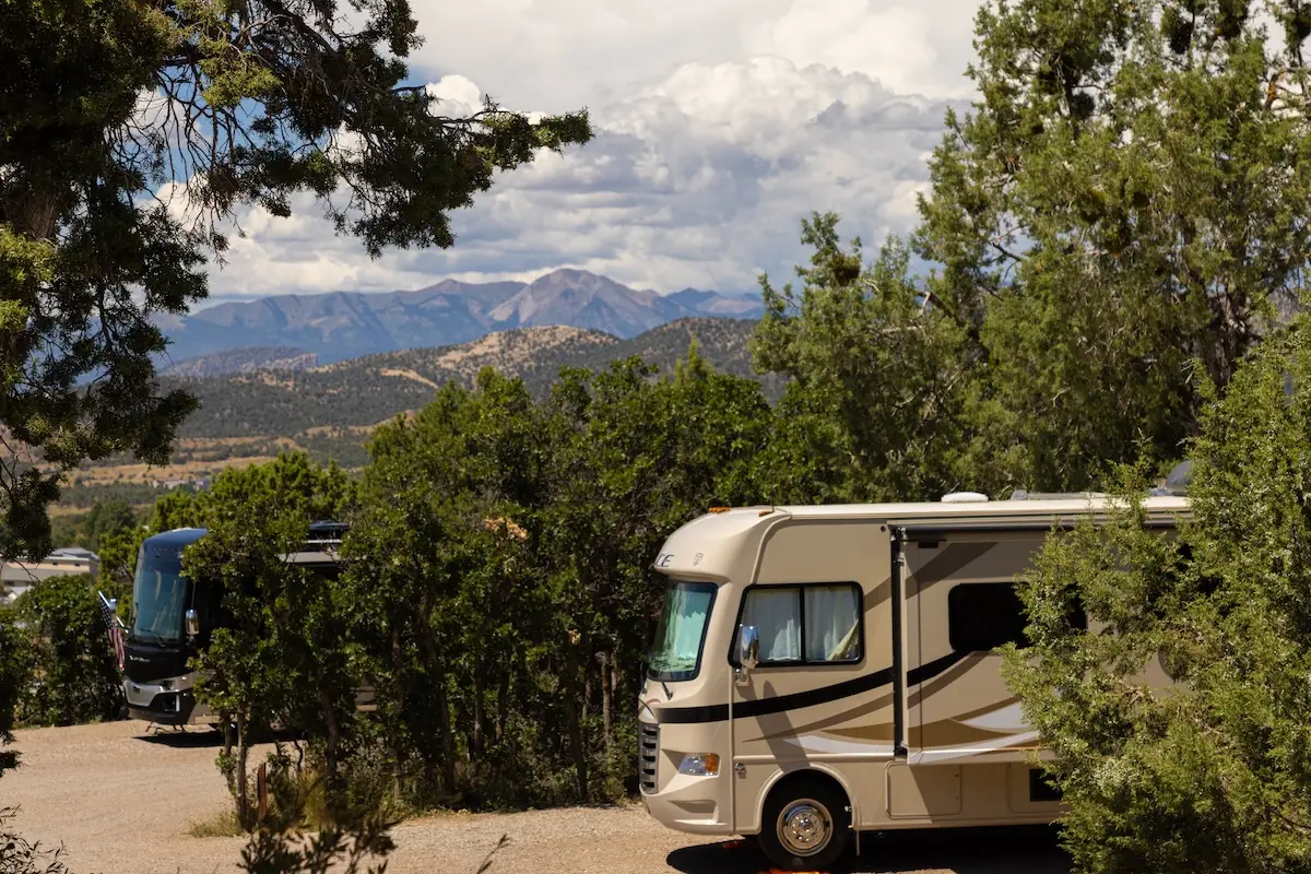 Two RVs parked among trees at the Oasis Durango Resort with mountains in the background under a cloudy sky.