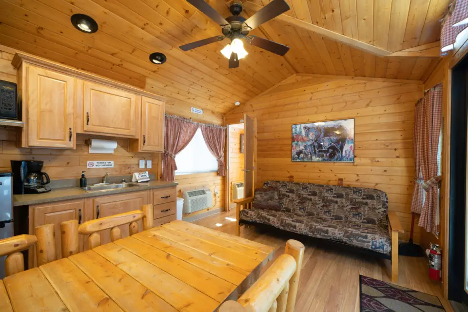 The cozy cabin interior at Oasis Durango Resort features wooden walls and ceiling. There's a dining table, chairs, sofa, kitchenette, and ceiling fan. A window with curtains and a painting decorate the space, creating an inviting atmosphere in one of our premium RV sites.