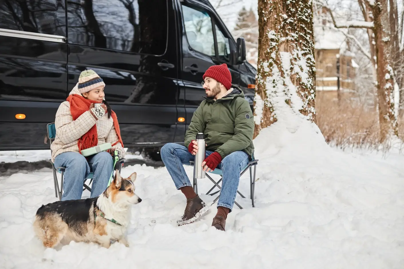 Two people sit on chairs in the snow next to a black van at Rayford Crossing, holding mugs. A dog stands nearby as they enjoy the crisp winter air in their cozy attire, ringing in the New Year with warmth and tranquility at this serene RV resort.