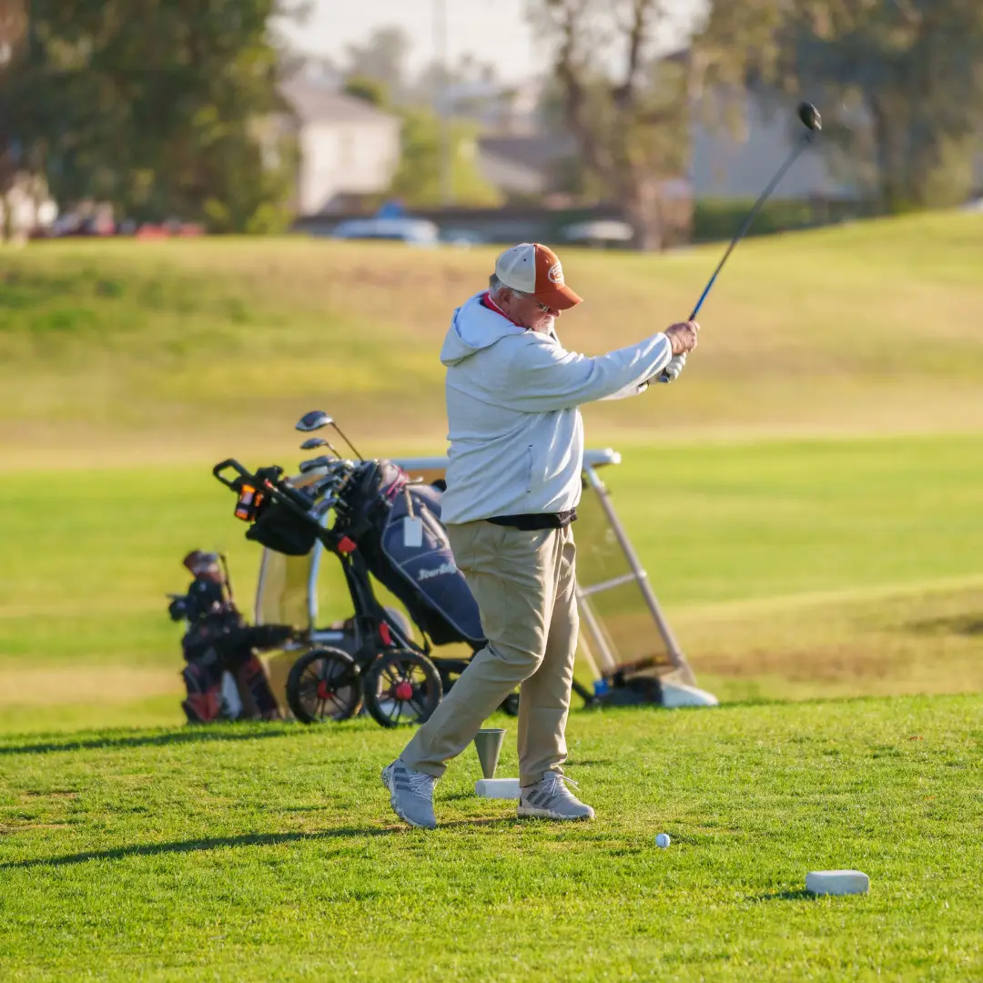 A golfer in a white jacket swings a club on a sunny AZ golf course. Golf bags are visible in the background, capturing the essence of spring adventures in El Mirage.