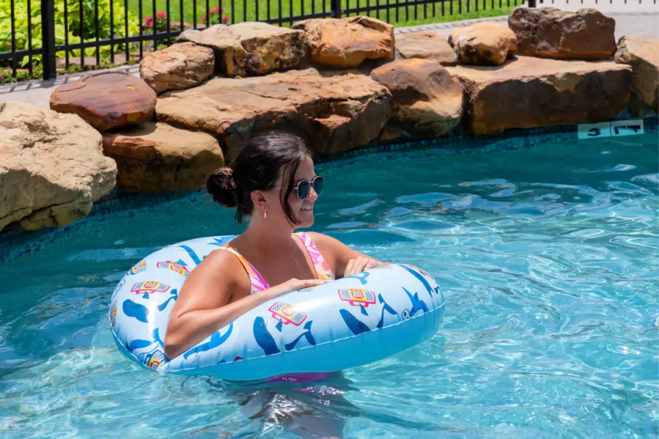 At Rayford Crossing in Spring, Texas, a person in sunglasses enjoys a leisurely float on a shark-themed inflatable ring in the sparkling swimming pool. Behind them, rocks add a touch of natural beauty to this idyllic setting perfect for monthly RV stays.