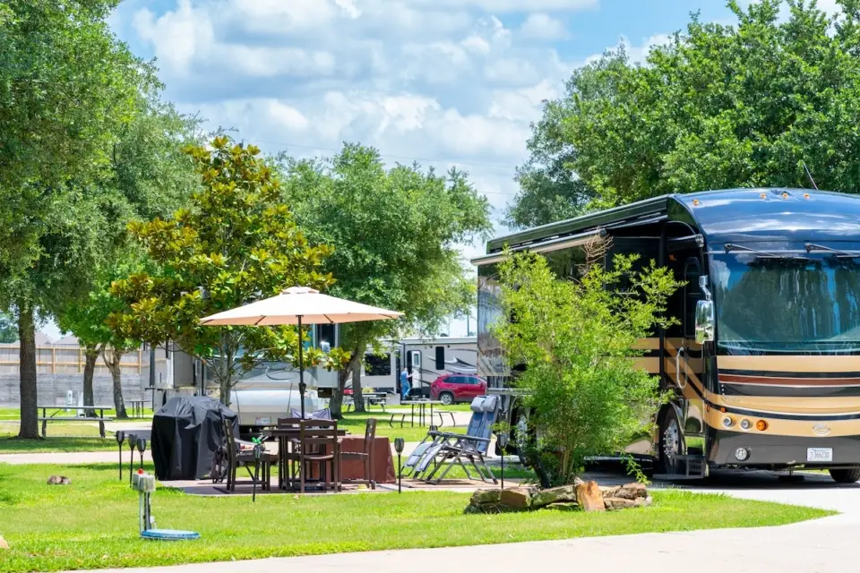 A large RV is parked at a scenic RV resort near a picnic area, complete with a table, chairs, and an umbrella under the sunny Spring Texas sky. Ideal for long monthly stays, this spot offers both relaxation and adventure.