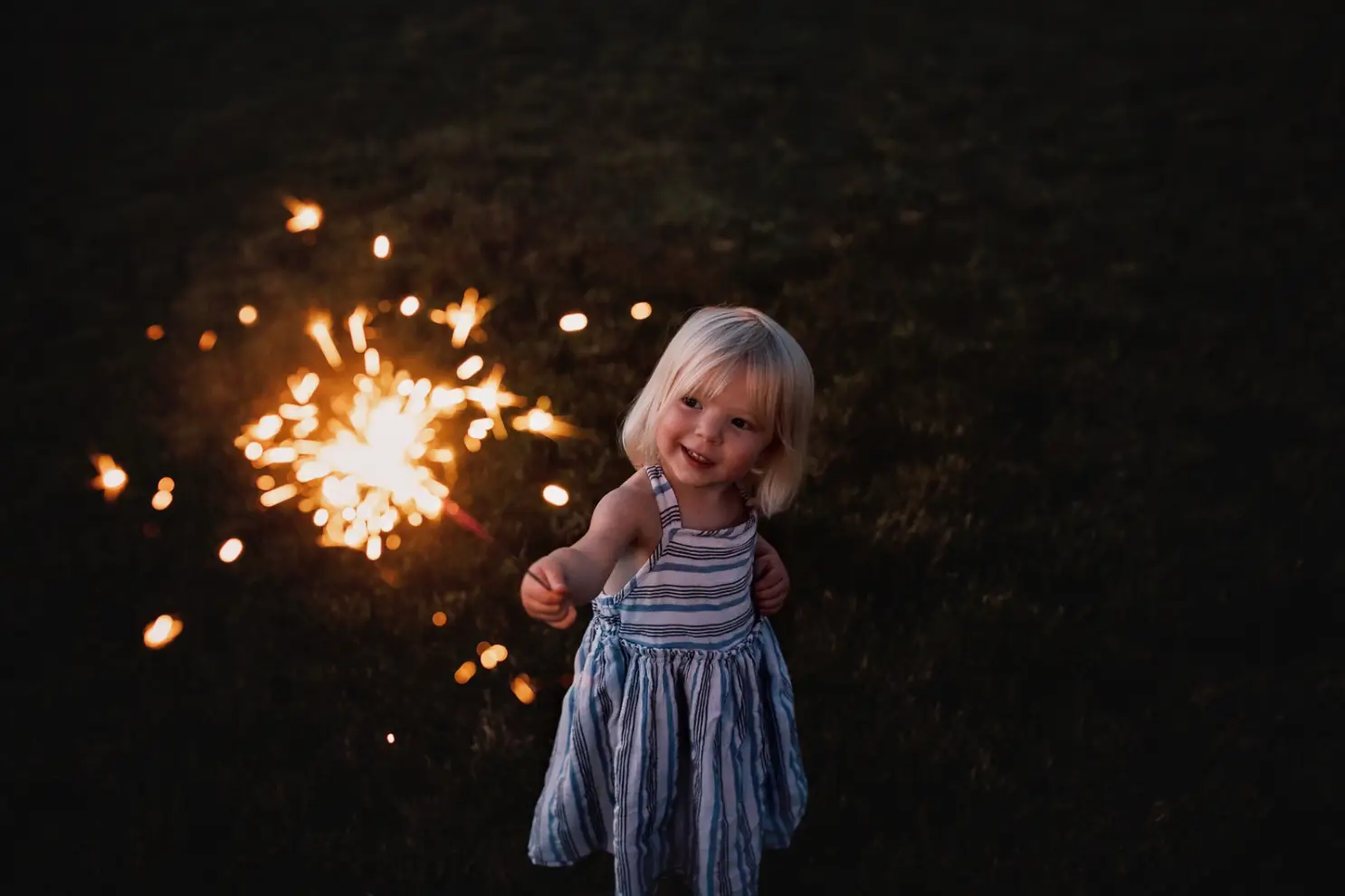 A young child with blonde hair holds a sparkling firework in the grassy, dimly lit area of Rayford Crossing RV Resort, smiling at the camera as holiday magic fills the air.