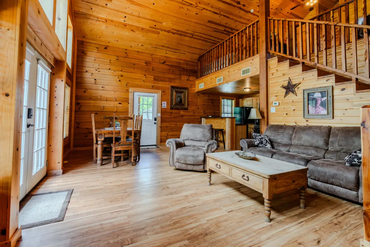 Rustic wooden cabin interior near Lake Conroe with a high ceiling, cozy seating area, staircase, and dining table. Perfect for holidays, the walls and floor are made of wood. Charming decor includes framed pictures and a star wall ornament.