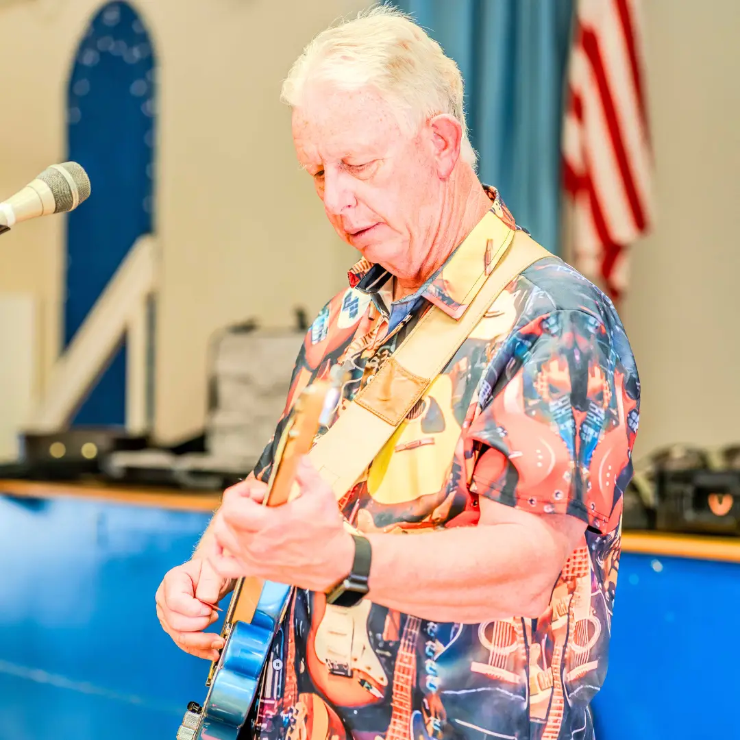Elderly man rocking an electric guitar on stage in Apache Junction, wearing a colorful guitar-patterned shirt, with the American flag proudly waving in the background.