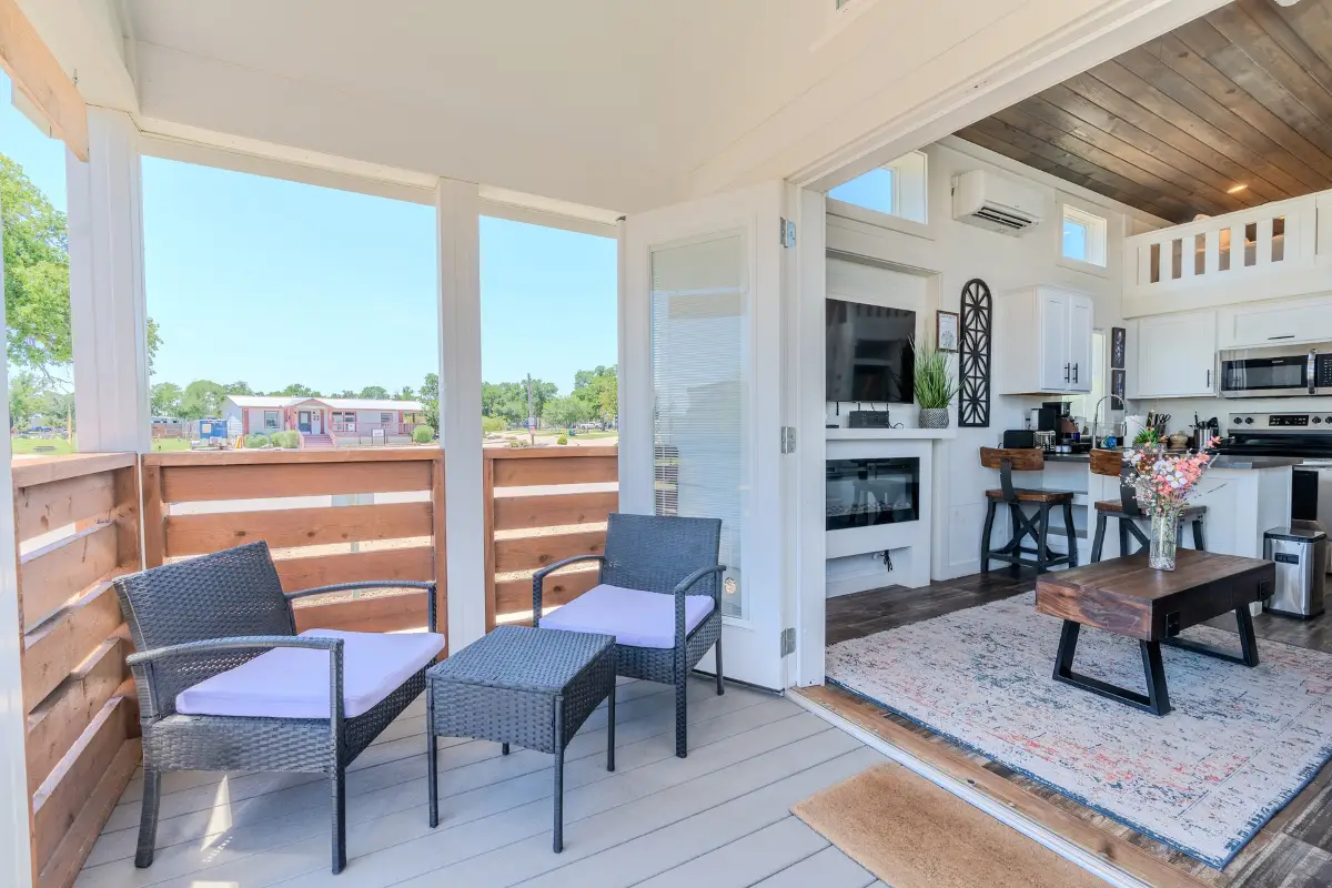 Modern open-plan tiny house interior at Oak Forest RV Resort, featuring a kitchen, living area, and loft. The adjacent deck has two wicker chairs with cushions. Large windows and doors flood the space with natural light, perfect for Thanksgiving getaways in nature's embrace.