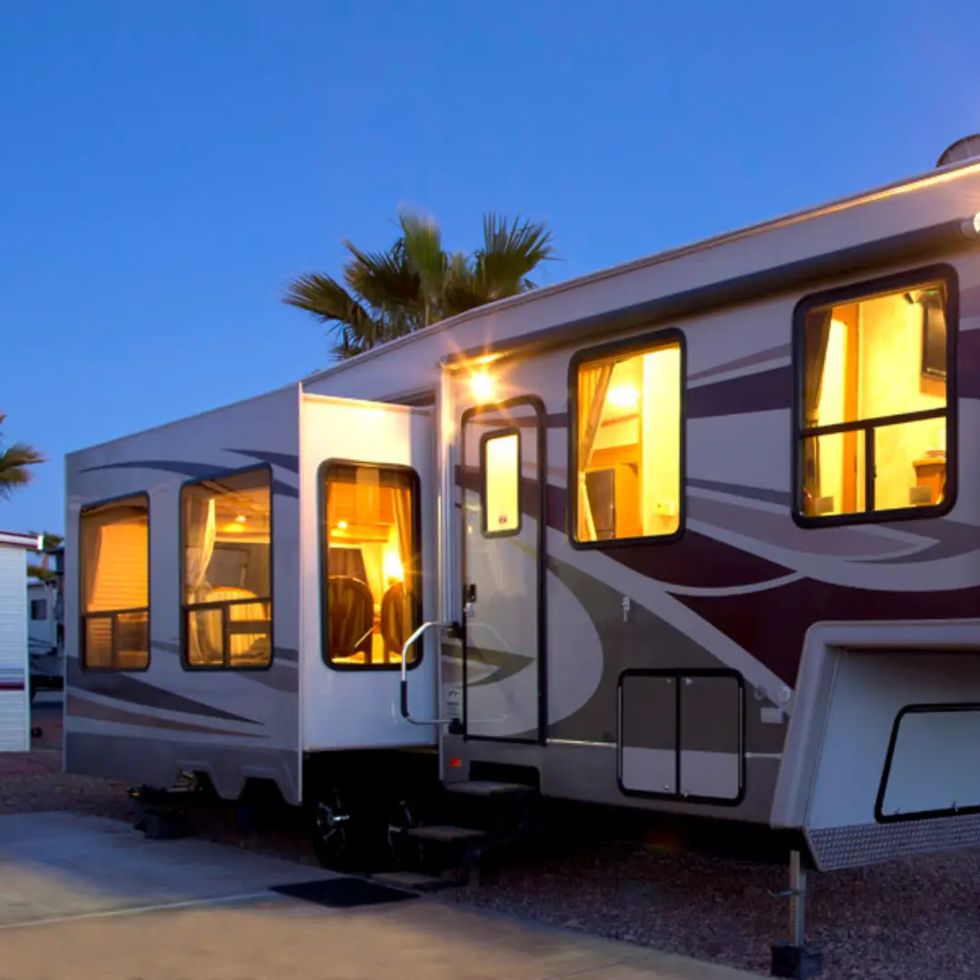 A modern RV with lit interior windows is parked on a gravel site at dusk, framed by Arizona's iconic palm trees. Perfect for early booking, this spot is a winter visitor's dream.