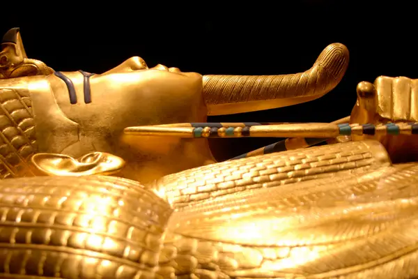 A close-up of a golden ancient Egyptian burial mask, featuring detailed carvings and inlaid colorful stones against a black background, as showcased in the science museum's exhibits.