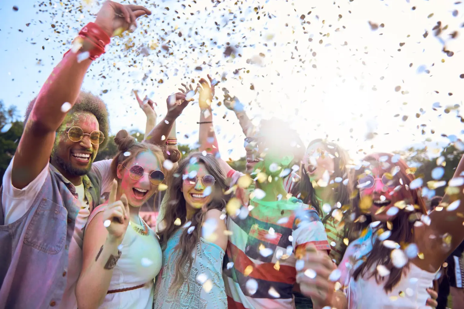 A group of people, like an auto draft of pure joy, celebrating outdoors in the sunlight, throwing colorful confetti and smiling amidst the trees in the background.