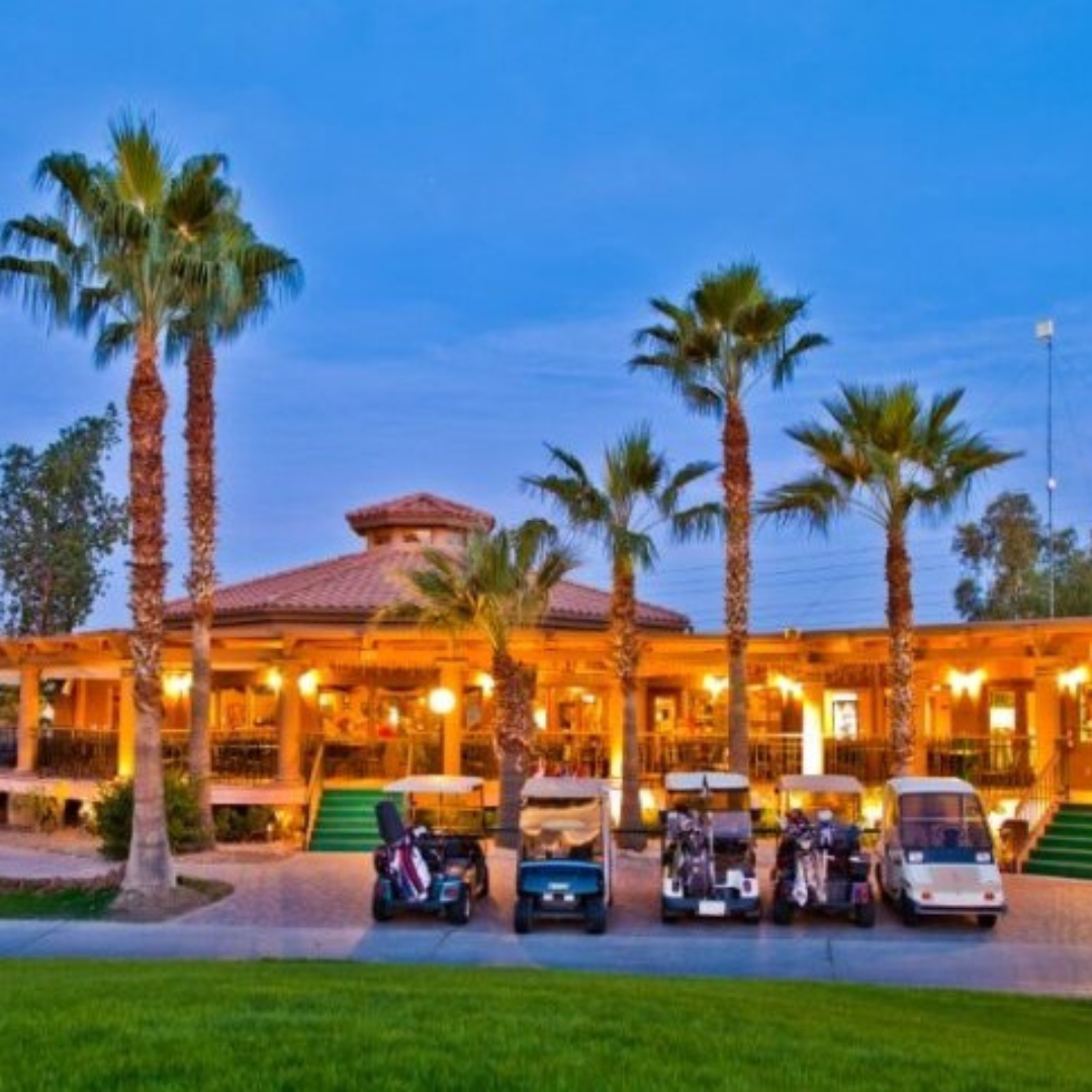 A clubhouse with a tiled roof is surrounded by palm trees, capturing the essence of Pueblo El Mirage. Several golf carts are parked in front on a paved area, ready for January activities. The sky is clear and blue.