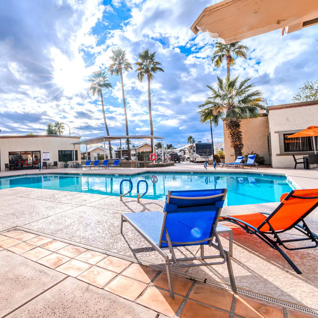 Experience the beauty of Arizona's winter at Sunrise RV Resort, where the outdoor swimming pool area is adorned with bright lounge chairs and palm trees under a clear blue sky with scattered clouds.