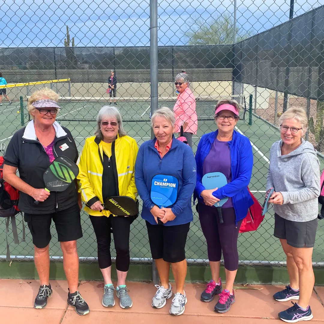 Five women stand on a pickleball court at Sunrise RV Resort, paddles in hand. They wear casual sportswear and smile at the camera, embracing their long-term stays. A chain-link fence and other players are visible in the background.