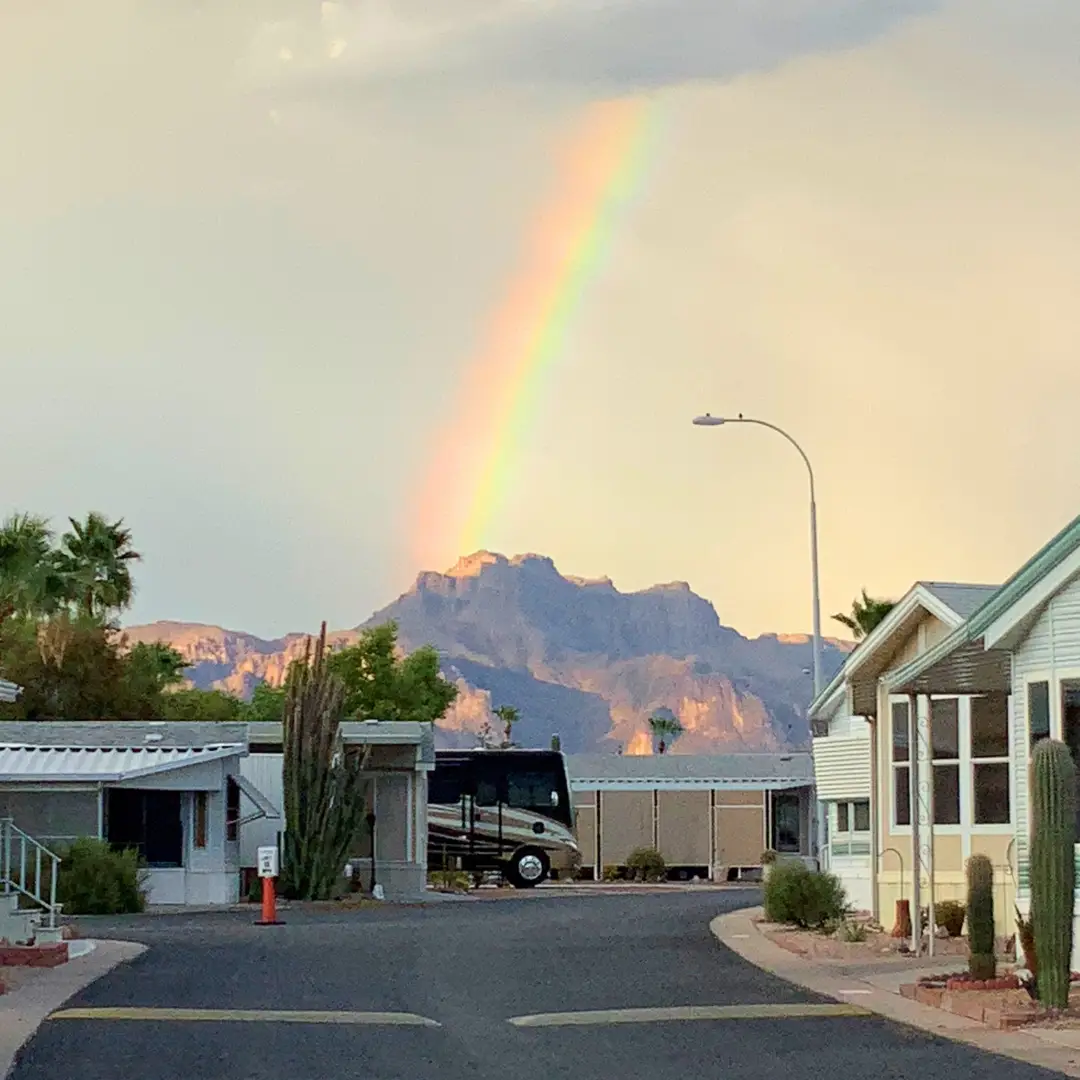 A vibrant rainbow arcs over the mountain landscape, viewed from Sunrise RV Resort, where mobile homes and cacti dot the foreground, perfect for long-term stays.