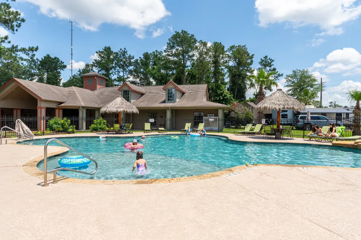 Children gleefully splash in the outdoor swimming pool at Rayford, surrounded by lounge chairs and a building with a thatched roof. The scene, set against a backdrop of lush trees and a clear blue sky, epitomizes summer programming fun for everyone.