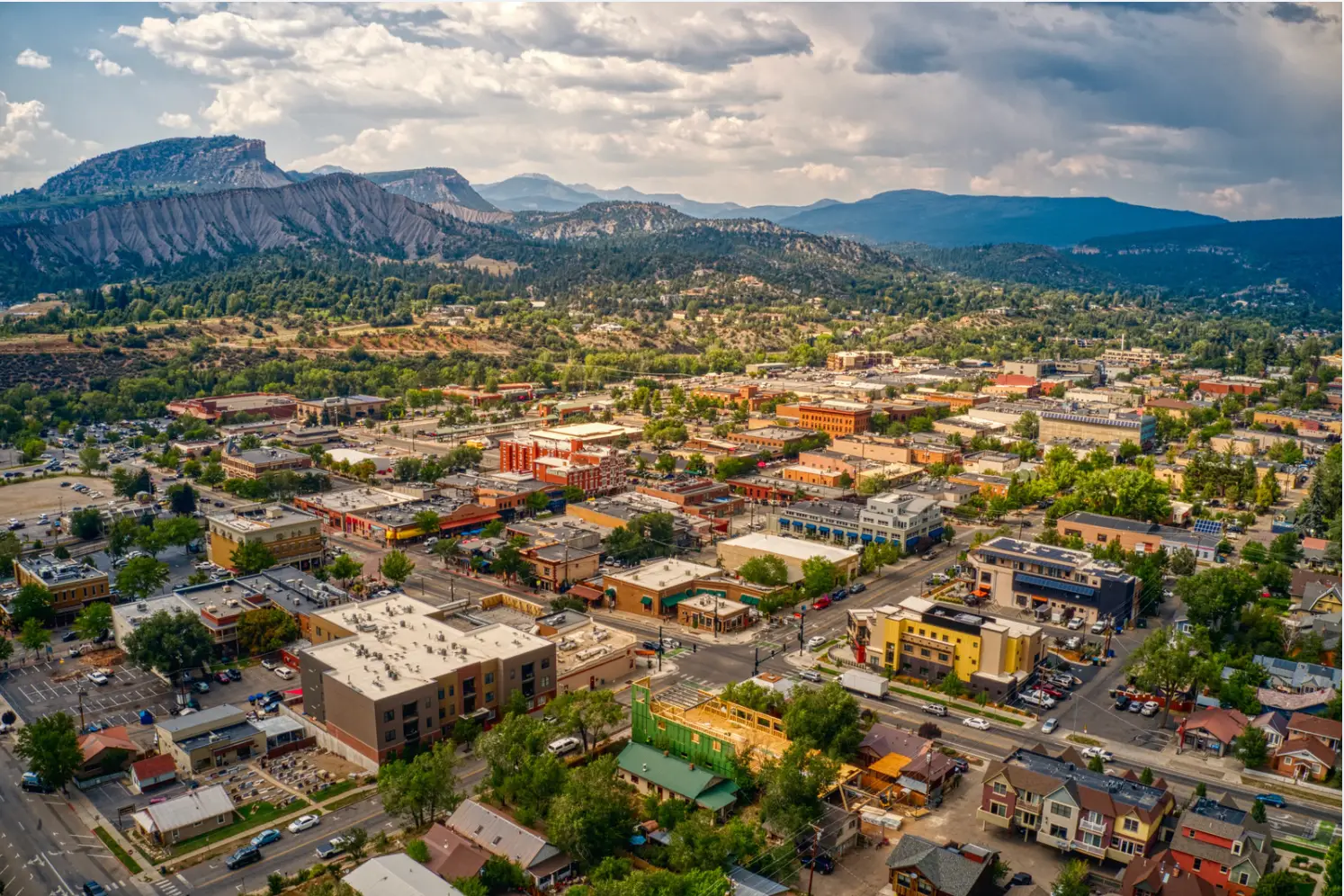 Aerial view of a small town with buildings, roads, and vehicles, surrounded by trees and mountains under a partly cloudy sky—truly the perfect time for an outdoor adventure.