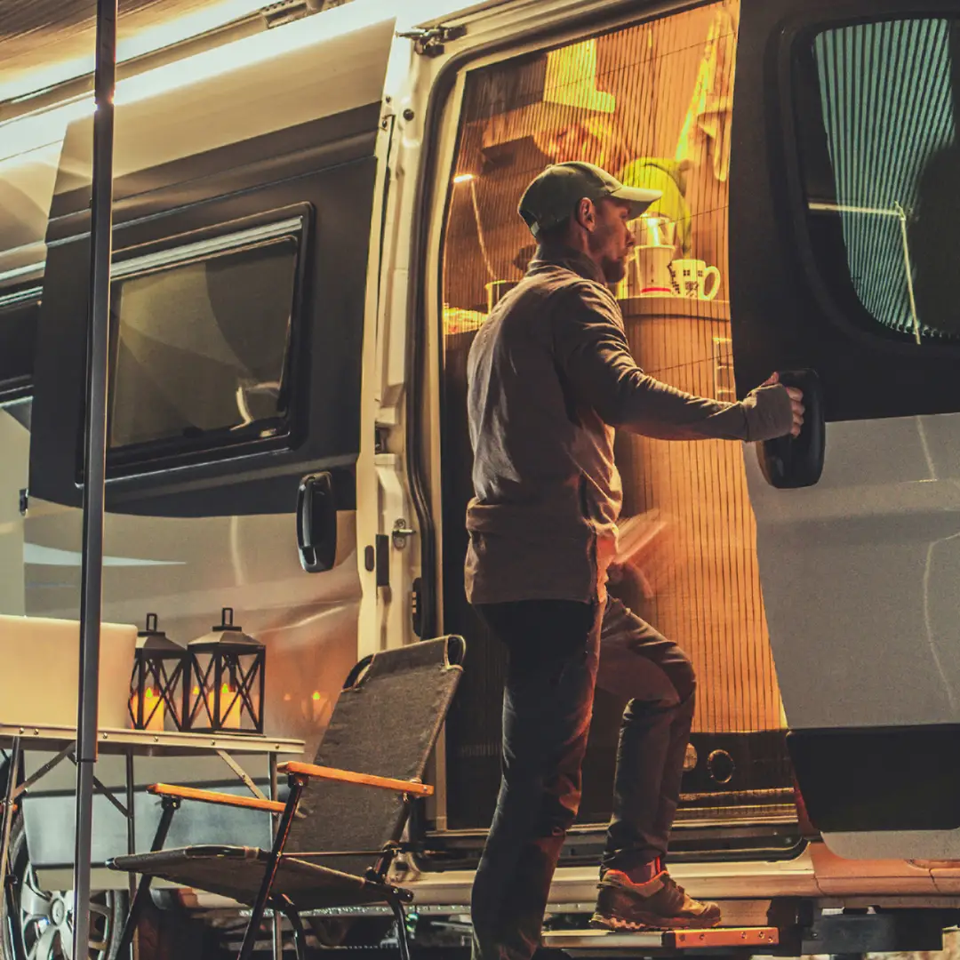 A person in casual clothing stands at the open door of a well-lit camper van interior, ready for a Spring and Summer Getaway. Just outside, a folding chair invites relaxation amidst the scenic beauty of Durango, Colorado.