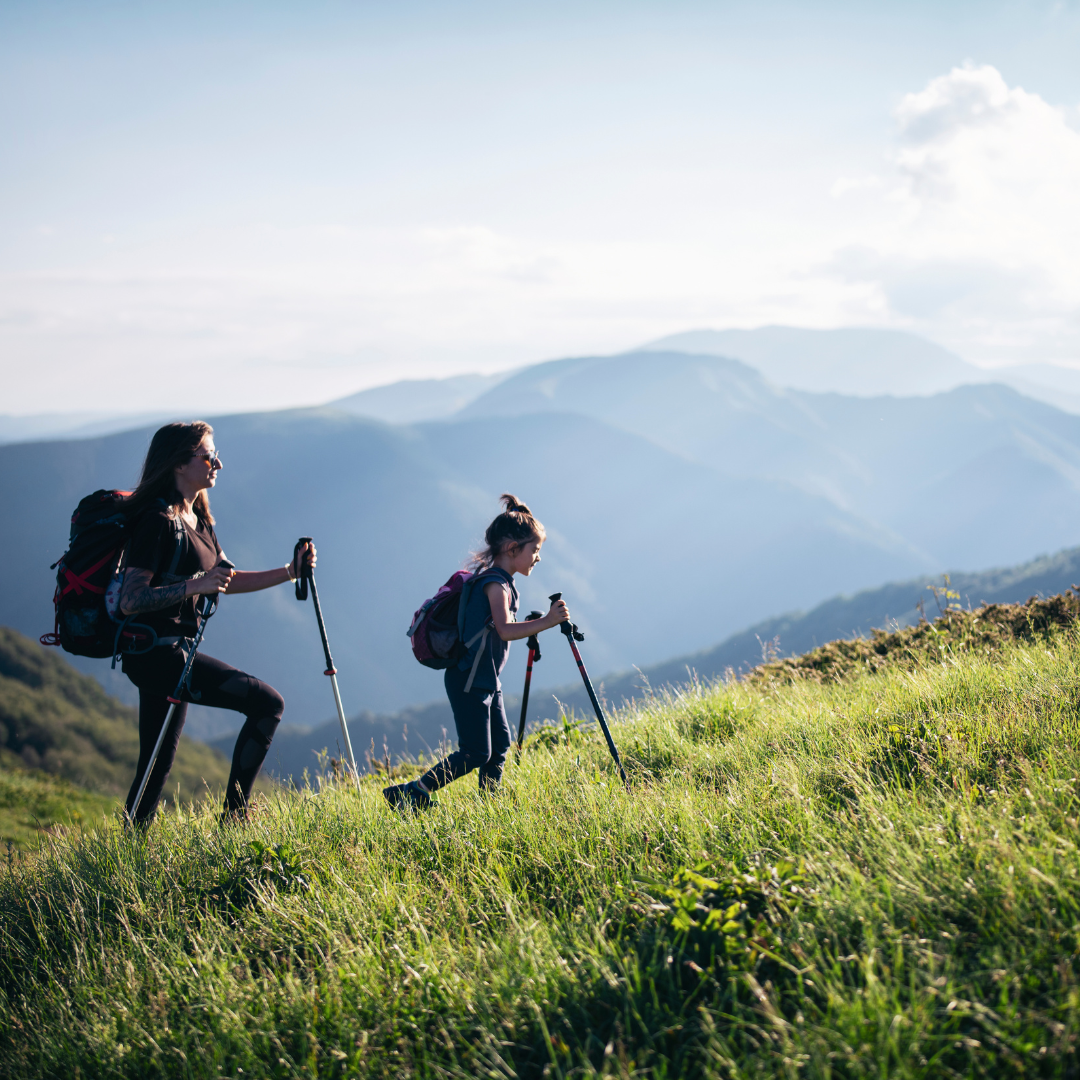 Two people with hiking poles walk uphill on a grassy mountain slope under a clear sky, enjoying their summer getaway, with the distant mountain ranges near Durango in the background.