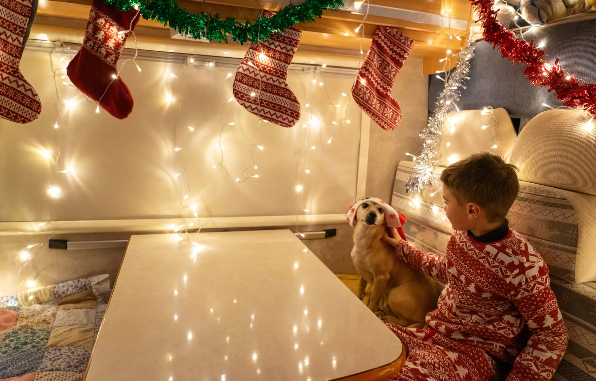 A child in red festive pajamas sits with a dog wearing reindeer antlers inside a decorated camper at Rayford Crossing, adorned with stockings and string lights—a perfect holiday getaway.