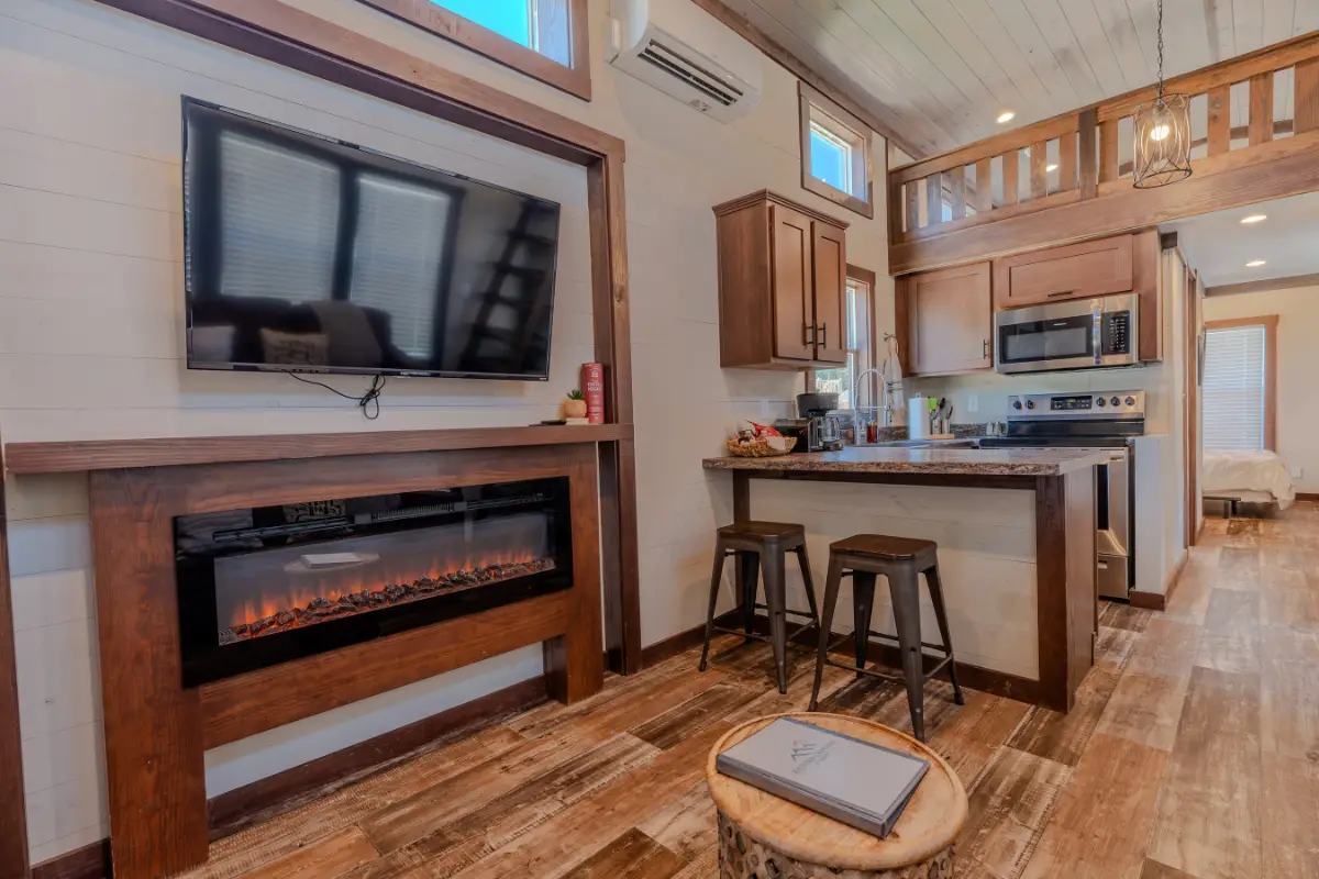 The interior of this compact wooden home at Rayford Crossing offers a perfect holiday getaway, featuring a cozy kitchen with stools, a wall-mounted TV, an electric fireplace, and a charming loft.