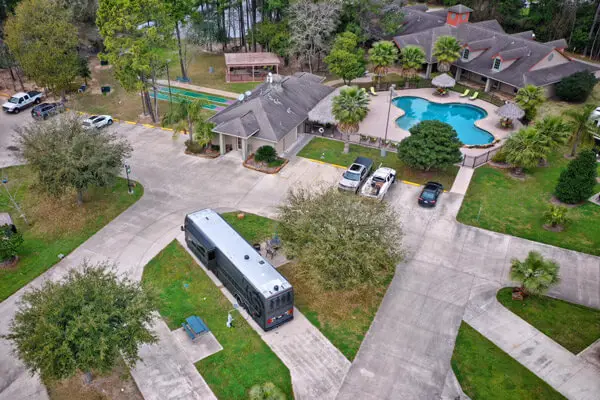 Aerial view of a residential area in Texas with a swimming pool, parked cars, and a bus amidst surrounding trees. Pathways connect buildings, offering a taste of Texas-sized fun for both locals and those looking to travel through this vibrant community.