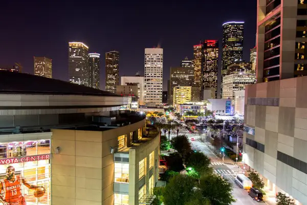 Night view of a vibrant cityscape in Texas, with illuminated skyscrapers and buildings. An arena is visible in the foreground, surrounded by bustling streets lined with streetlights, offering a glimpse of the fun and excitement that awaits travelers.