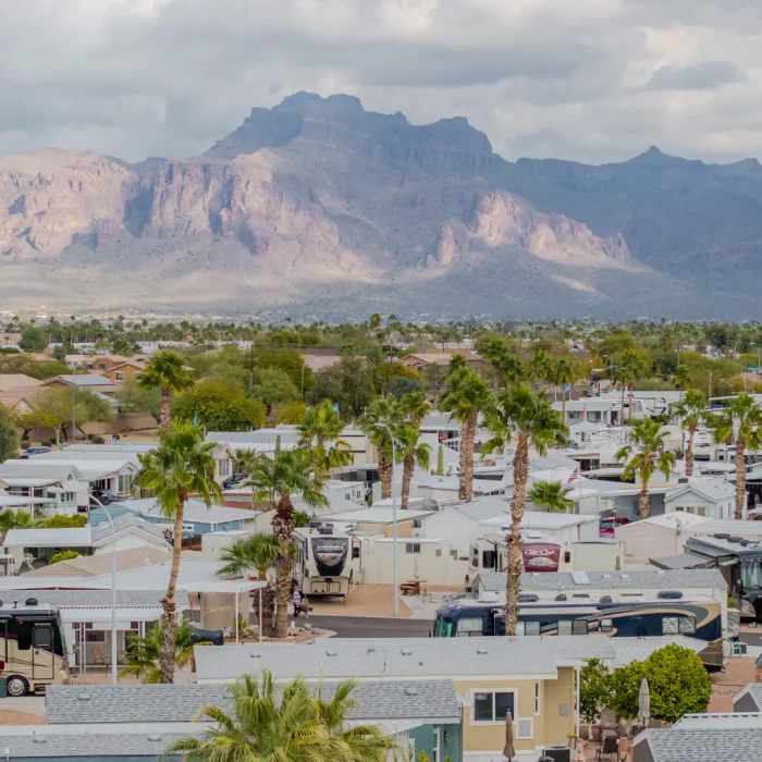 Aerial view of Sunrise RV Resort, a trailer park community nestled among palm trees with a majestic mountain range in the background. The landscape captures Arizona winter beauty beneath a cloudy sky.