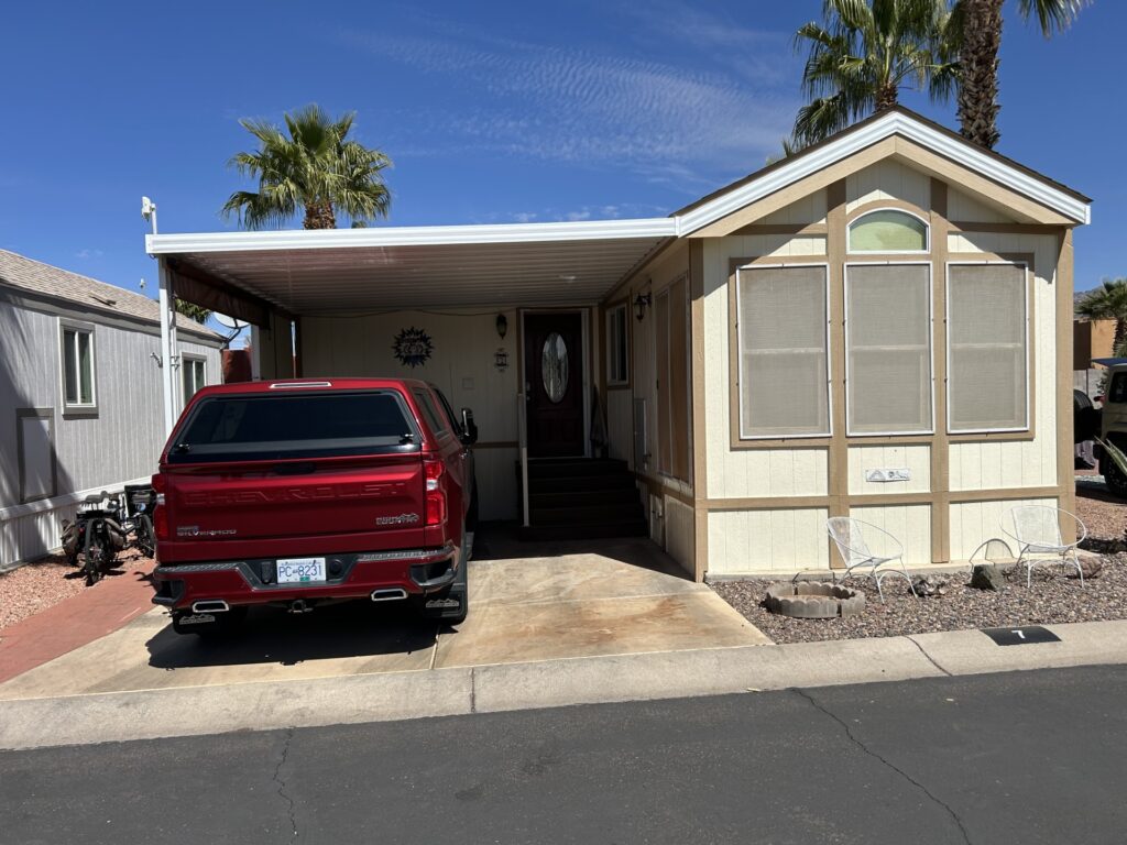 A beige modular home with an attached porch and steps is next to a parked red truck. The yard has gravel and palm trees under a clear blue sky.