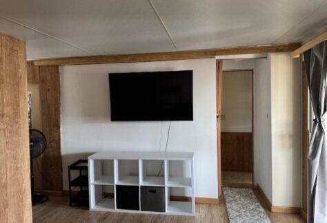 A wall-mounted flat-screen TV above a white cubby shelf with storage bins in a room with wood accents and a floor fan in the corner.