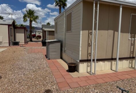 A beige mobile home with a small storage shed, red paver walkway, and gravel landscaping under a partly cloudy sky with palm trees in the background.