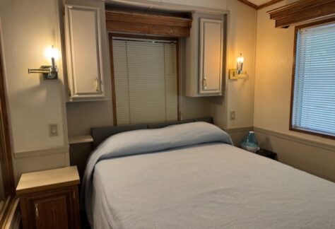 A neatly made bed with a light blue cover is centered between two built-in cabinets and wall sconces, next to windows with closed blinds in a small, tidy bedroom.