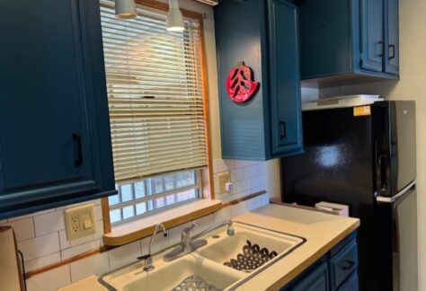 A kitchen sink with a dish rack, blue cabinets, a window with blinds above the sink, and a black refrigerator to the right.