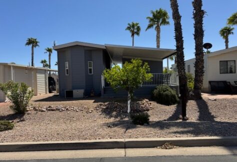 A single-story gray manufactured home with a covered porch sits in a desert landscape yard with rocks, small bushes, and palm trees under a clear blue sky.