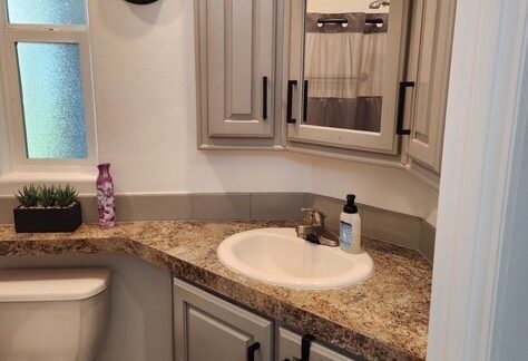 Small bathroom with beige cabinets, a granite countertop, a white sink, a wall mirror, a clock, a window, a potted plant, a soap dispenser, and folded towels hanging on a rack.