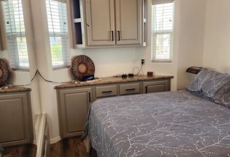 A small bedroom with a bed covered in a gray patterned comforter, a built-in cabinet, two windows, and a decorative plate on the counter.