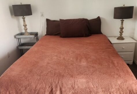 A bed with a rust-colored bedspread and brown pillows is centered between two nightstands, each with a lamp, in a plain, white-walled room.