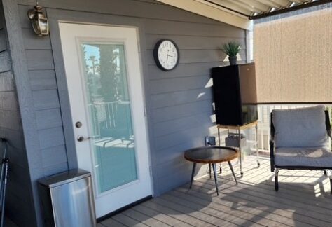 A covered patio with gray siding, a glass door, a wall clock, a trash can, a small round table, a chair, and a TV on a stand with a plant on top.