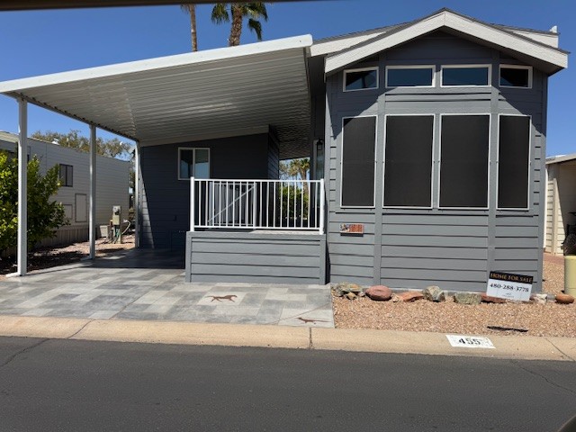 Gray manufactured home with a covered porch, paved driveway, and a "For Sale" sign in front, located in a desert landscaping setting.
