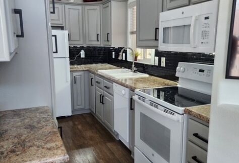 Narrow kitchen with gray cabinets, white appliances, brown countertops, dark tile backsplash, wood flooring, and decorative vases on the upper cabinets.
