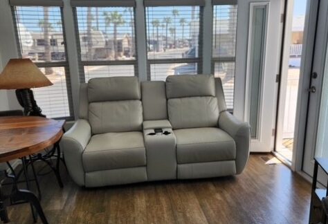 Beige recliner sofa with cup holder centered in a living area, wood floor, round table with lamp on left, large windows and open door letting in daylight.