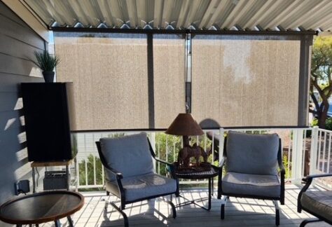 A covered patio with three cushioned chairs, a small round table, a lamp on a side table, vertical blinds, and a potted plant in sunlight.