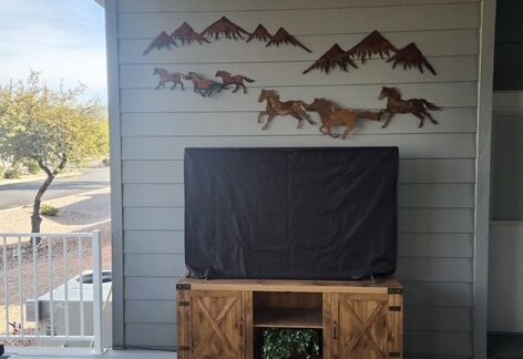 A covered TV sits on a wooden stand against a gray paneled wall with metal horse and mountain decorations above. A rug and partial porch railing are also visible.