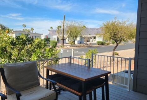 A small patio with a chair, table, and bench overlooks a suburban street lined with desert landscaping and palm trees under a blue sky.
