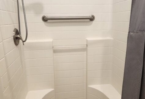 White tiled shower with a handheld showerhead, grab bar, two built-in corner seats, a shower curtain, and a textured mat on the floor.