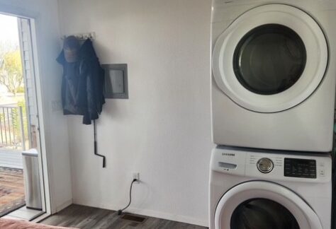 A stacked washer and dryer are next to a bed in a room with wood flooring and an open door leading outside. A coat rack with jackets is mounted on the wall.
