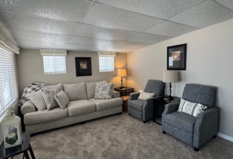 A living room with a beige sofa, two gray armchairs, a lamp, framed wall art, and carpeted flooring. Light enters through windows with blinds and valances.