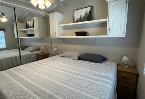 A neatly made bed with striped bedding is centered between two nightstands, under white shelves and cabinets, with a ceiling fan and mirrored closet doors visible.