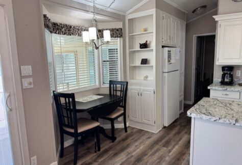 A small dining nook with a black table and two chairs is set by a window. The kitchen features white cabinets, granite countertops, a refrigerator, and hardwood floors.