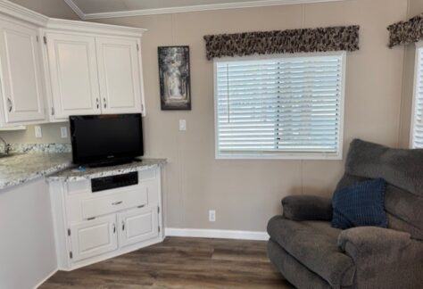 Living room corner with white cabinets, a small TV, a window with blinds and valance, a gray recliner chair with a blue pillow, and wood-style flooring.