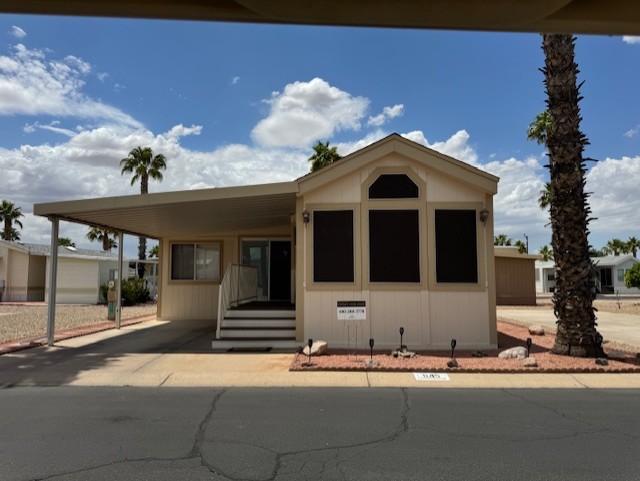A beige manufactured home with a covered carport, front steps, and black window screens, set in a desert neighborhood with palm trees and gravel landscaping.