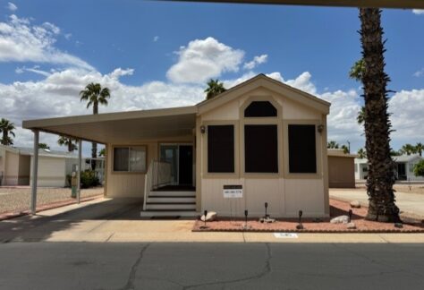 A beige manufactured home with a covered carport, front porch, and landscaped yard with rocks and palm trees, set in a sunny residential area under a partly cloudy sky.